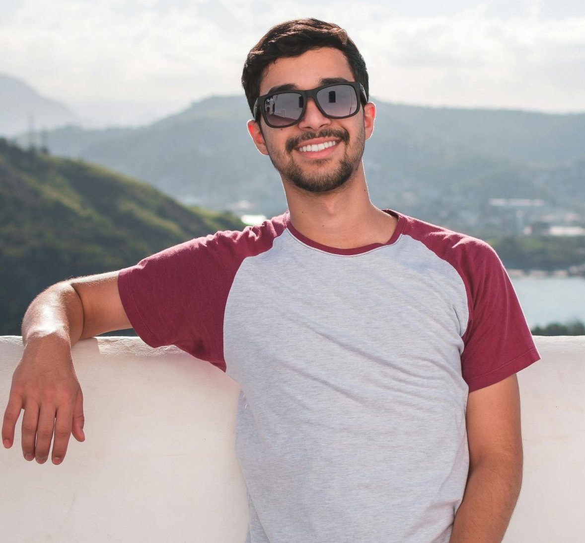 Young man in sunglasses leaning on a balcony with a scenic city view in the background.