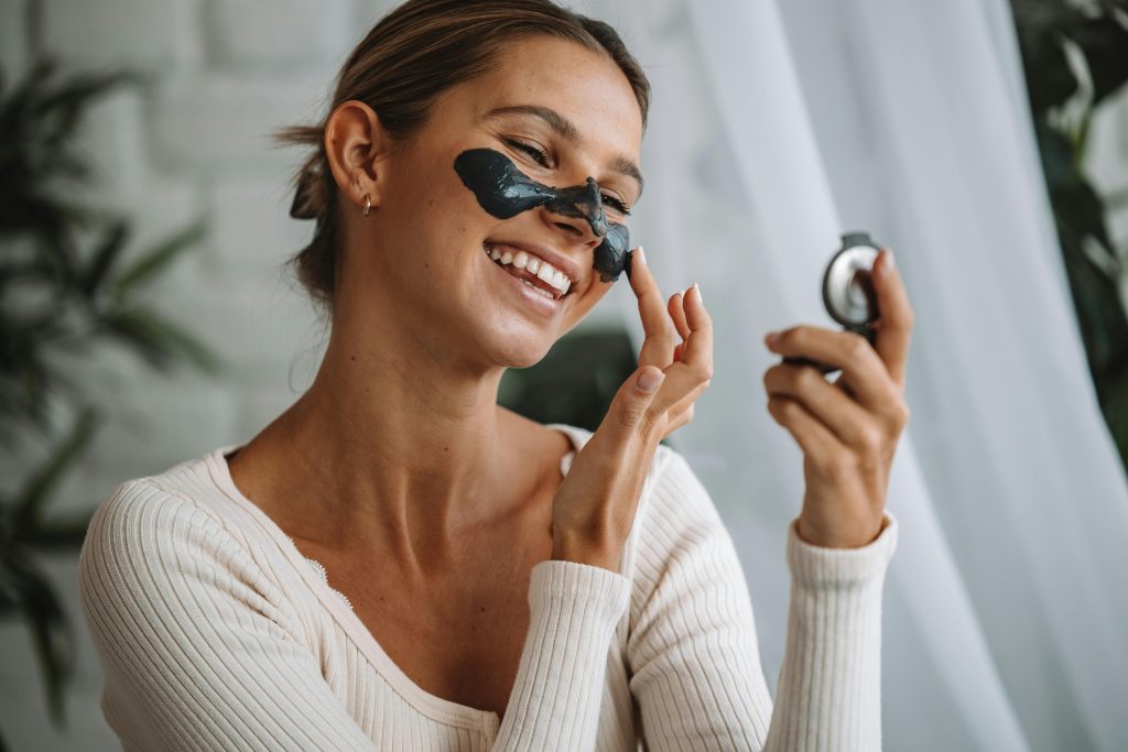 Smiling woman applying a cosmetic face mask while looking in a mirror indoors.