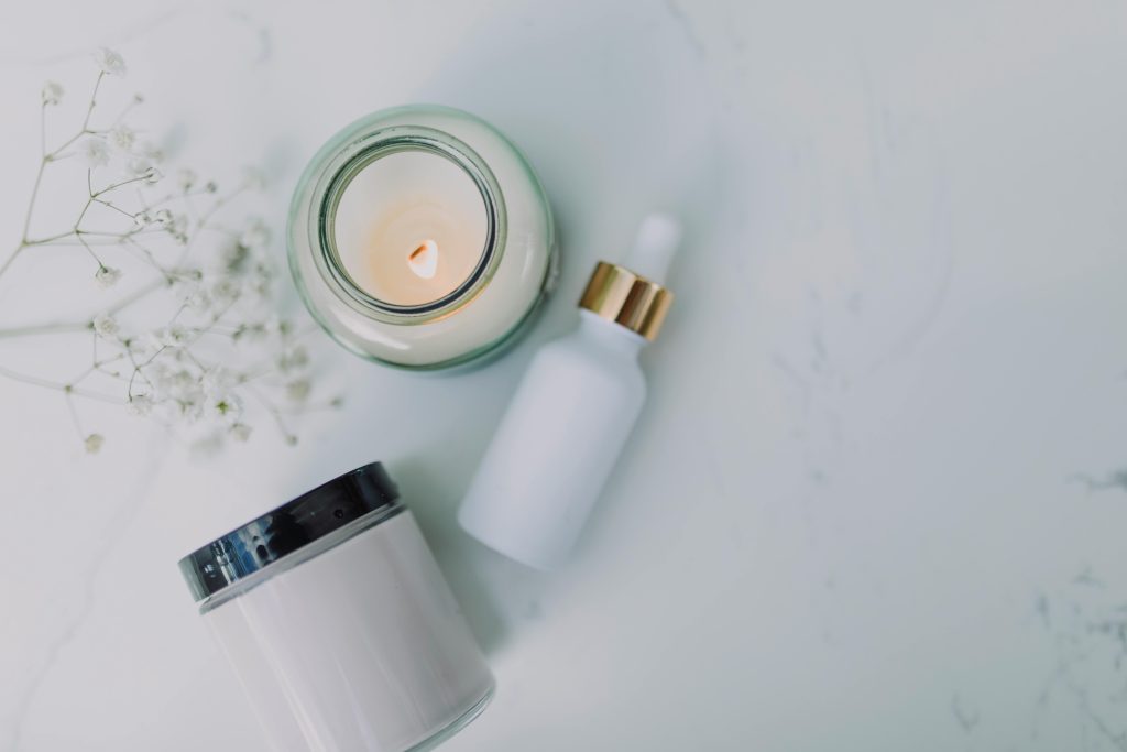Overhead view of spa essentials including a candle, dropper bottle, and jar on a white marble surface.