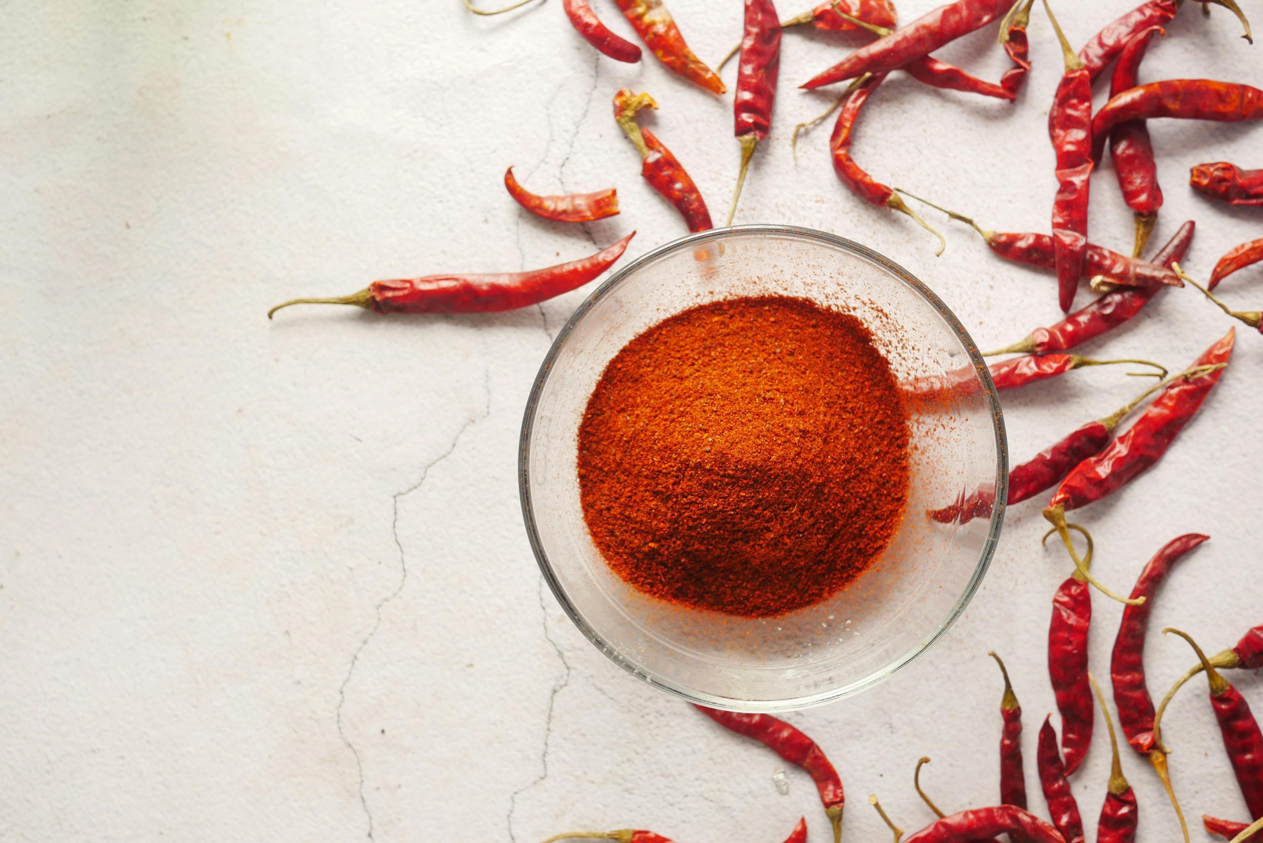 pexels-photo-8693381-8693381 Bowl of vibrant chili powder surrounded by red chili peppers on a textured white background.