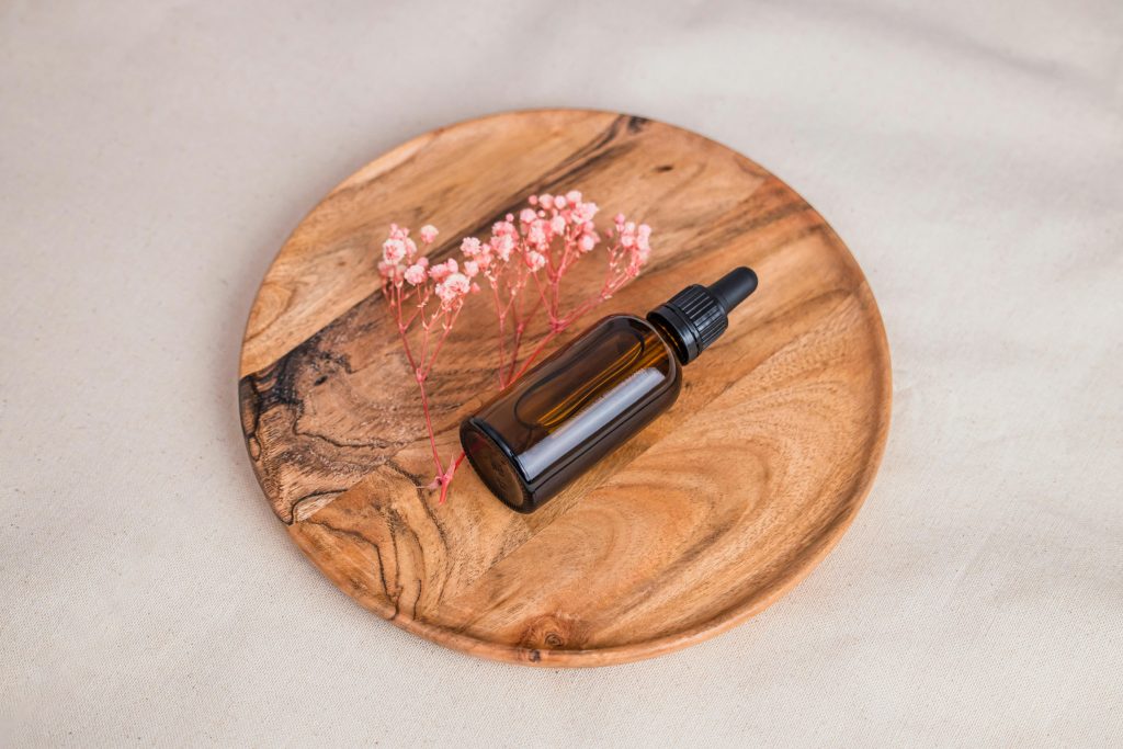 Elegant glass dropper bottle on a wooden tray with pink flowers, ideal for skincare and beauty concepts.