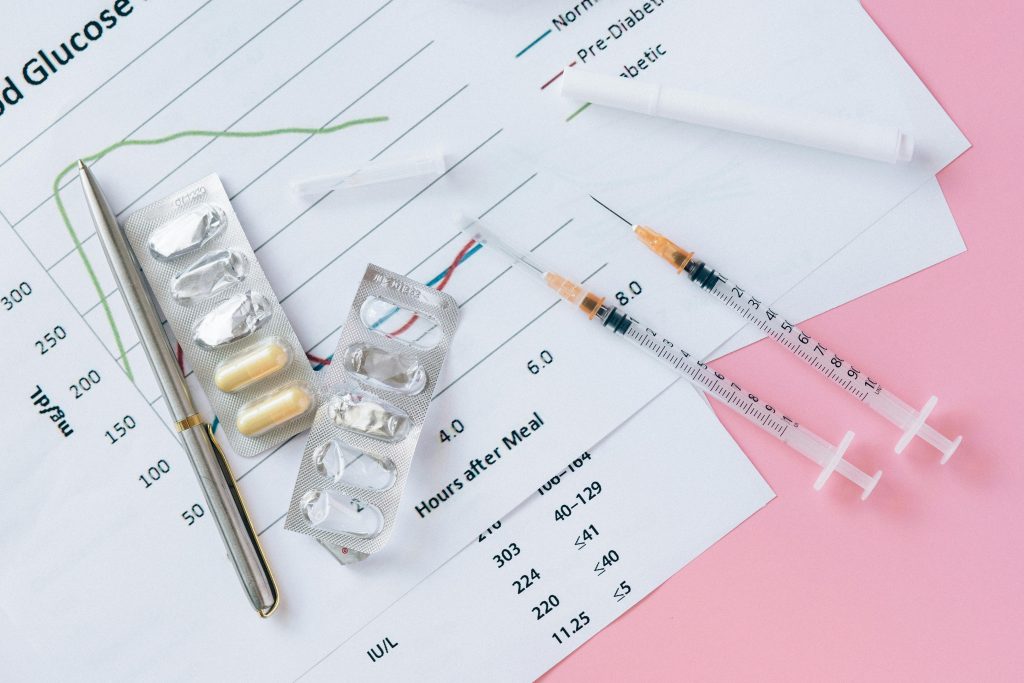 Flat lay of medicines, syringes, and a blood glucose chart on a pink surface, representing health management.